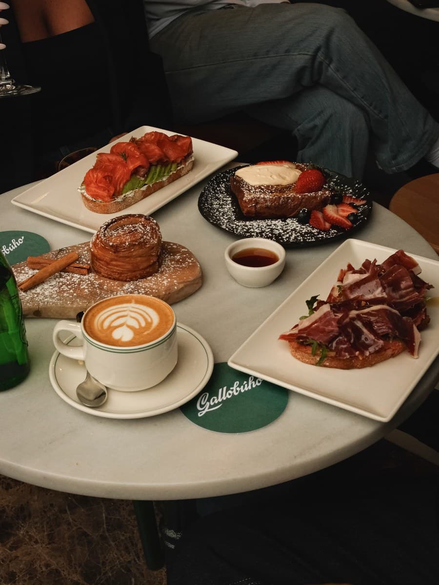 Gallobúho brunch: salmon avocado toast, French toast with strawberries, cinnamon-dusted croissant, jamón ibérico on bread, cappuccino with latte art, and house coasters on a marble table.